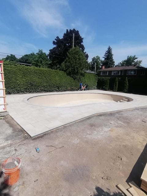 Empty concrete pool under construction in a backyard with hedges and trees in background