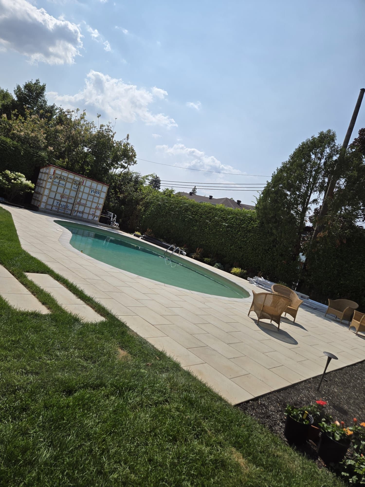 Backyard with rectangular swimming pool on tiled patio, surrounded by green lawn and trees under partly cloudy sky