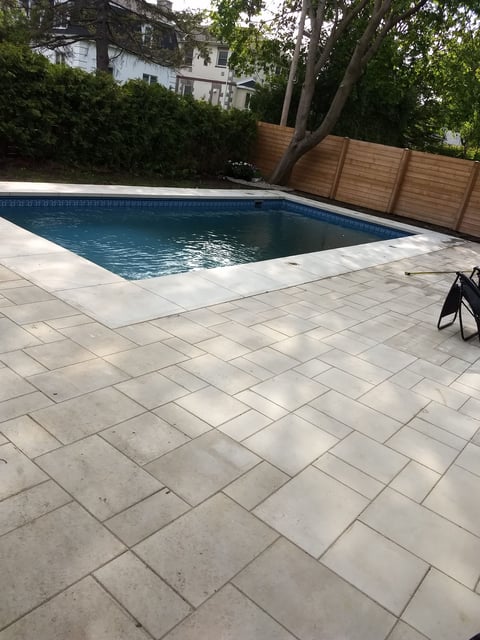 Rectangular backyard swimming pool with blue water on light-colored paved deck, surrounded by wooden fence and green trees, with residential building visible in background