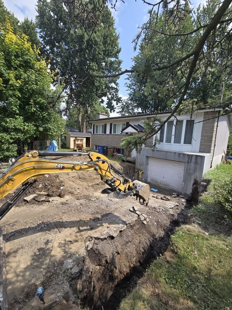 Yellow excavator on residential lot next to house under construction surrounded by tall trees